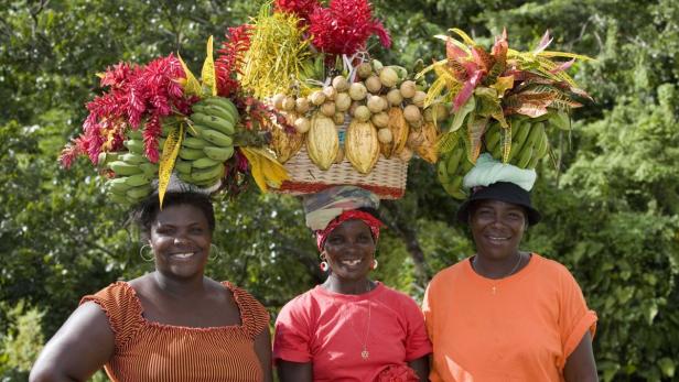 Drei Frauen tragen Körbe mit Bananen, Kakao und Blumen auf ihren Köpfen.