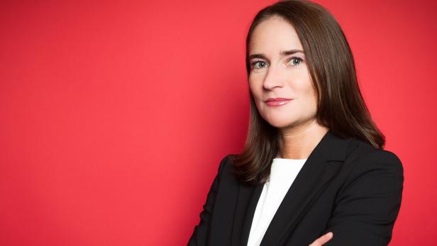 Eine Frau mit braunen Haaren und Blazer vor einem roten Hintergrund.