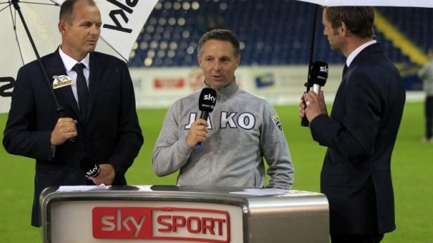 Drei Sky Sport Reporter stehen mit Mikrofonen im Regen vor einem Fußballstadion.