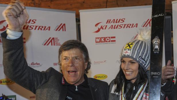 Ein Mann mit erhobener Faust und eine Frau mit Skiern posieren vor einem „Haus Ski Austria“-Banner.