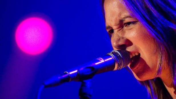Eine Sängerin singt mit Leidenschaft in ein Mikrofon vor blauem Hintergrund mit einem pinkfarbenen Scheinwerfer.