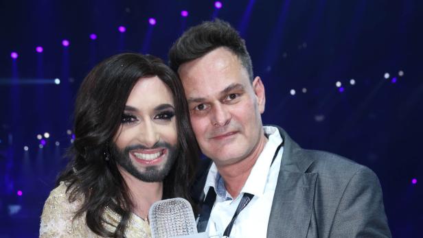 Conchita Wurst posiert mit einem Mann vor einem dunklen Hintergrund mit Lichtern.
