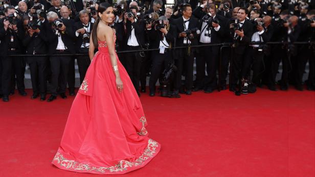 Freida Pinto in einem roten Kleid auf einem roten Teppich, umgeben von Fotografen.