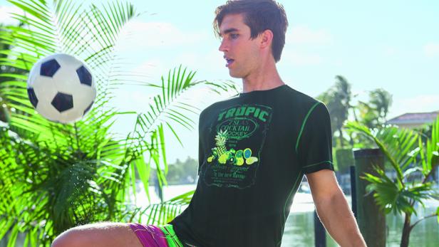 Ein junger Mann spielt mit einem Fußball vor tropischer Kulisse.