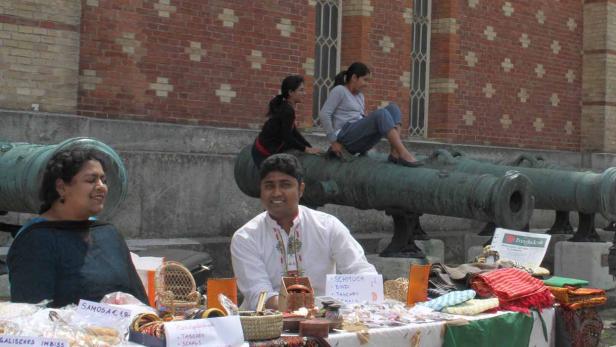 Ein Mann und eine Frau verkaufen bangladeschische Produkte an einem Stand, während zwei Frauen auf Kanonen sitzen.