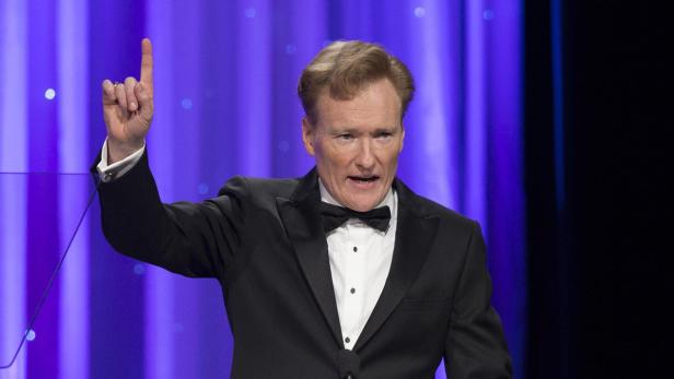 Conan O'Brien in einem schwarzen Smoking gestikuliert mit erhobenem Zeigefinger.