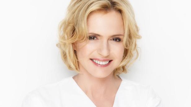 Eine lächelnde Frau mit blonden, gewellten Haaren und einem weißen Oberteil vor einem weißen Hintergrund.