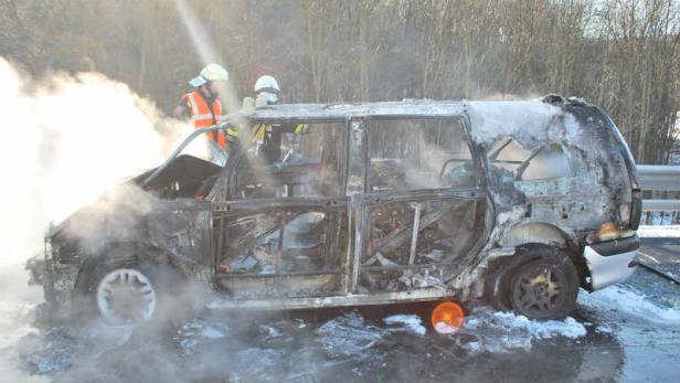 Ein ausgebranntes Auto steht auf der Straße, während Feuerwehrleute im Hintergrund arbeiten.