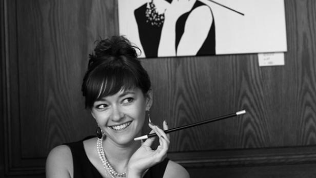 Eine Frau im Stil von Audrey Hepburn sitzt mit einer Zigarettenspitze in einem Café.