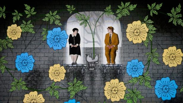 Eine stilisierte Bühnenszene mit zwei Darstellern vor einer bemalten Mauer mit Blumen und Blättern.