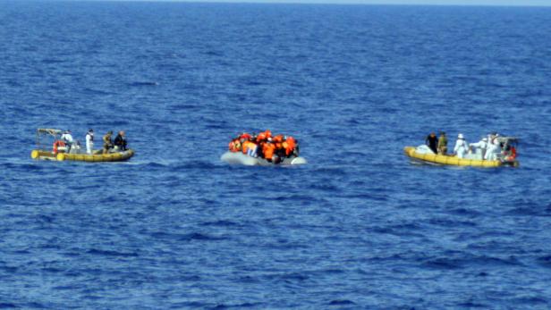 Drei Boote sind auf dem offenen Meer zu sehen, eines davon überfüllt mit Menschen in orangefarbenen Westen.