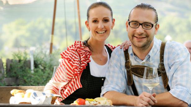 Ein Paar in Tracht sitzt an einem Tisch mit Brot, Wurst und Wein.