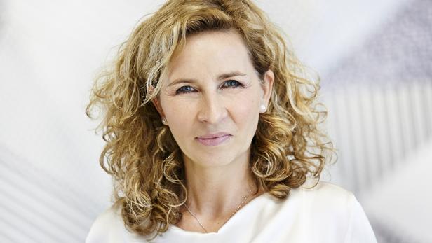 Porträt einer Frau mit blonden, lockigen Haaren und einem weißen Oberteil.