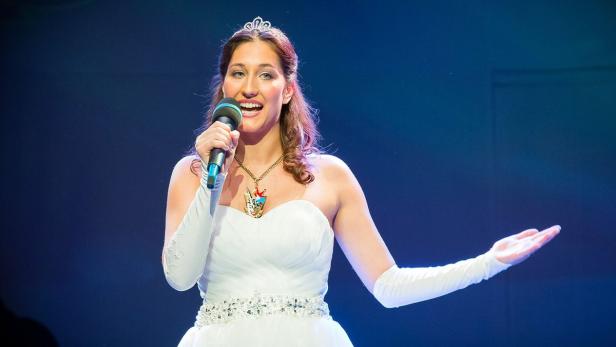 Eine junge Frau mit Tiara und weißem Kleid singt in ein Mikrofon.