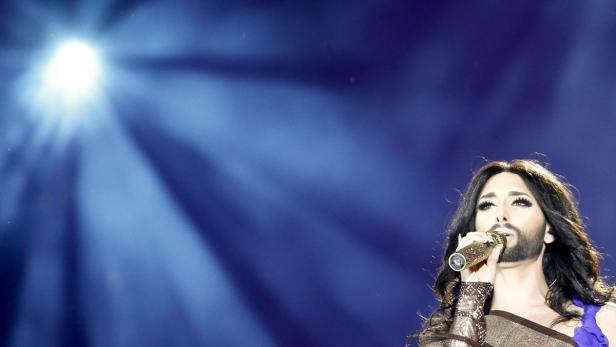 Conchita Wurst singt vor einem blauen Hintergrund mit Scheinwerfern.
