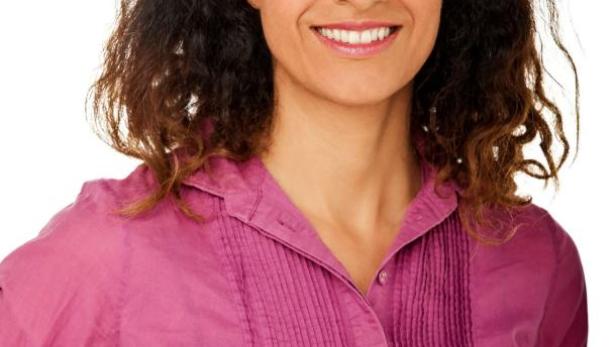 Eine lächelnde Frau mit lockigem Haar und einem pinkfarbenen Hemd vor einem weißen Hintergrund.