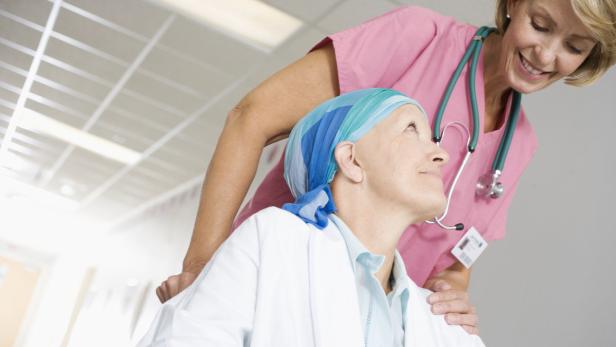 Eine Krankenschwester schiebt eine Patientin mit Kopftuch im Rollstuhl.