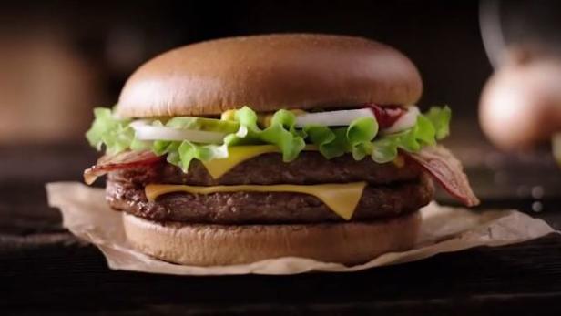 Ein doppelter Cheeseburger mit Speck, Salat, Zwiebeln und Soße auf einem Holztisch.