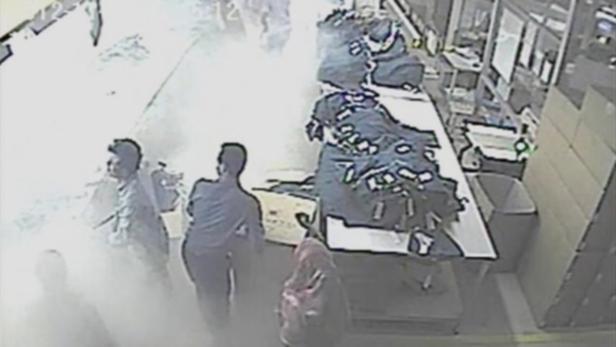 Überwachungsaufnahme zeigt Personen in einem Lagerraum mit Tischen und Waren.