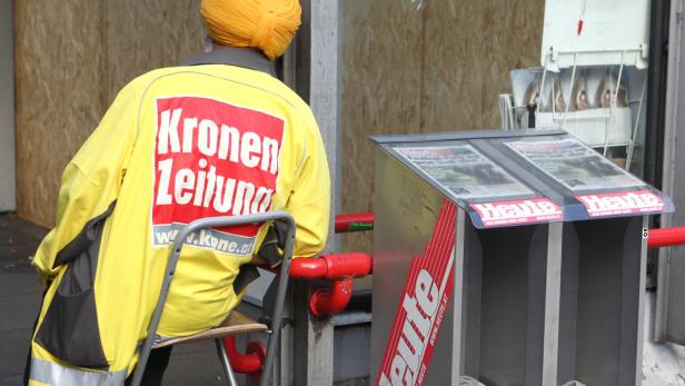 Ein Mann mit Turban in Arbeitskleidung der „Kronen Zeitung“ sitzt neben einem „Heute“-Zeitungsstand.