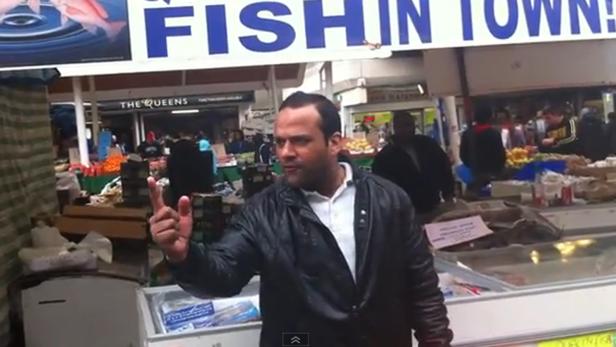 Ein Mann steht vor einem Fischstand auf einem Markt und gestikuliert.