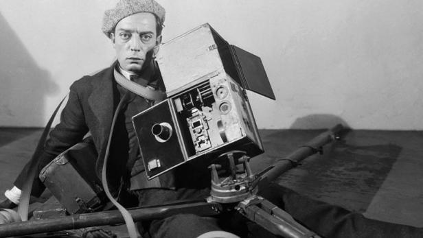 Der Schauspieler Buster Keaton sitzt mit einer alten Filmkamera auf dem Boden.