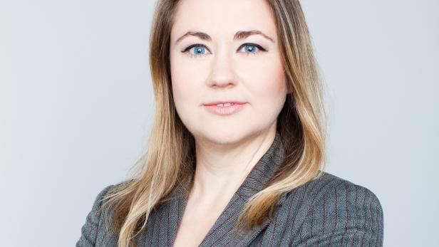 Eine Frau mit blonden Haaren und blauem Blazer vor einem hellgrauen Hintergrund.