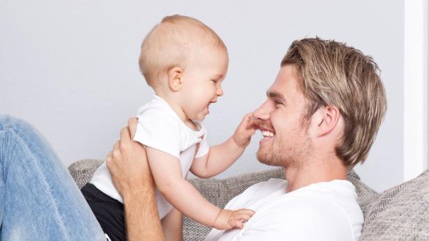 Ein lachender Vater liegt auf dem Sofa, während sein Baby seine Nase berührt.