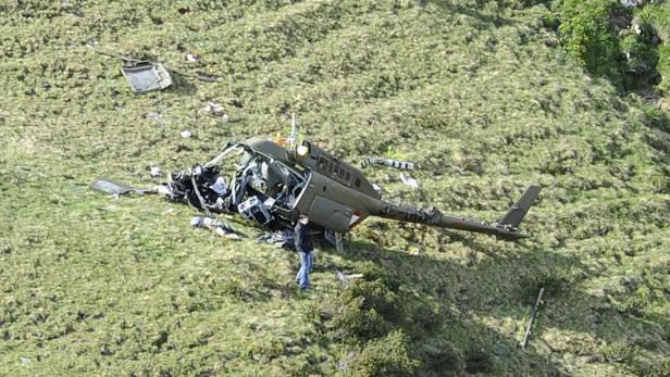 Ein abgestürzter Hubschrauber liegt auf einem grasbewachsenen Hügel, eine Person steht daneben.