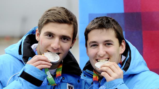 Zwei junge Männer mit blauen Jacken beißen in ihre Silbermedaillen.