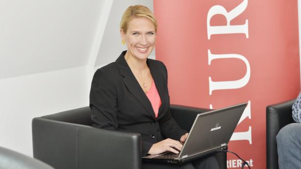 Eine blonde Frau sitzt in einem Sessel und arbeitet an einem Laptop.