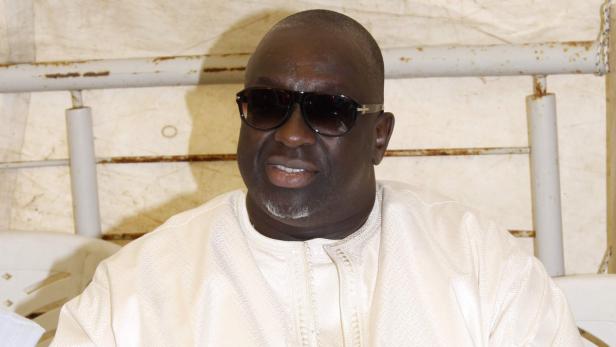 Ein Mann mit Sonnenbrille und hellem Hemd sitzt vor einem beigen Hintergrund.