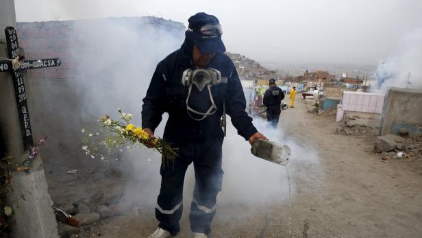 Ein Mann in Schutzkleidung gießt Wasser auf einen Friedhof, während er Blumen hält.