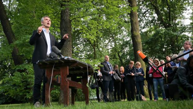 Ein Mann in Anzug spricht vor Mikrofonen im Freien zu einer Gruppe von Menschen.