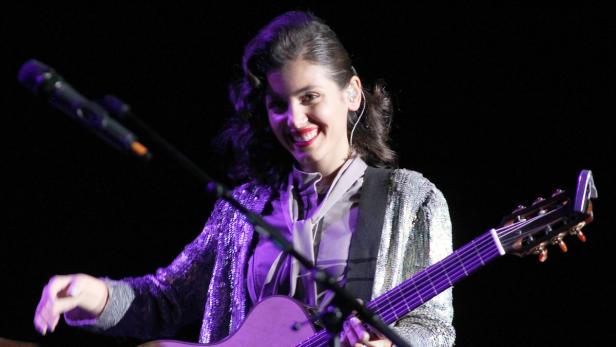 Eine lächelnde Frau mit Gitarre und glitzerndem Blazer auf einer Bühne.