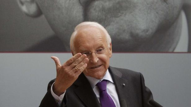 Ein älterer Mann mit Brille gestikuliert vor einem grauen Hintergrund.