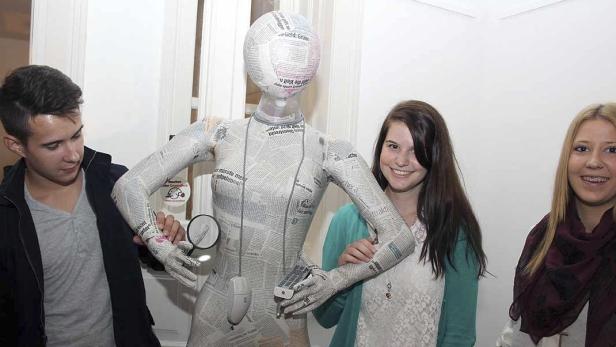 Ein junger Mann betrachtet eine mit Zeitungspapier beklebte Schaufensterpuppe mit einer Lupe, während zwei junge Frauen lächeln.