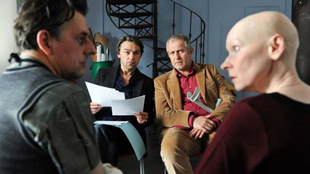 Vier Personen sitzen in einem Raum, zwei Männer halten Papiere, eine Frau hat eine Gehhilfe.