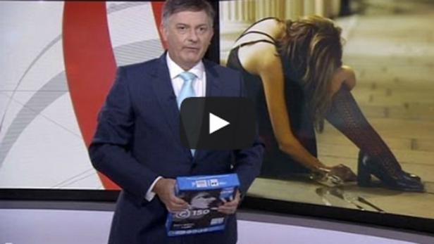 Ein Nachrichtensprecher hält eine blaue Schachtel vor einem Bildschirm, der eine Frau in Not zeigt.