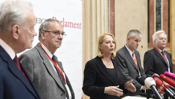 Eine Frau spricht bei einer Pressekonferenz vor Mikrofonen, umgeben von anderen Personen.