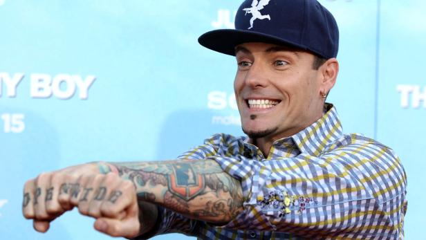 Ein lächelnder Mann mit Tattoos und einer Kappe posiert vor einer blauen Wand.
