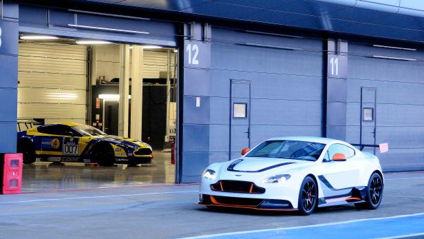 Ein weißer Aston Martin steht vor einer Garage, ein gelb-blauer Rennwagen ist im Hintergrund zu sehen.