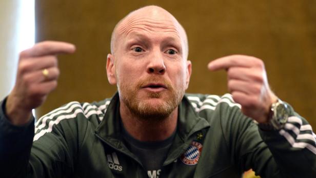 Ein Mann mit Bart und Trainingsjacke gestikuliert mit beiden Händen.