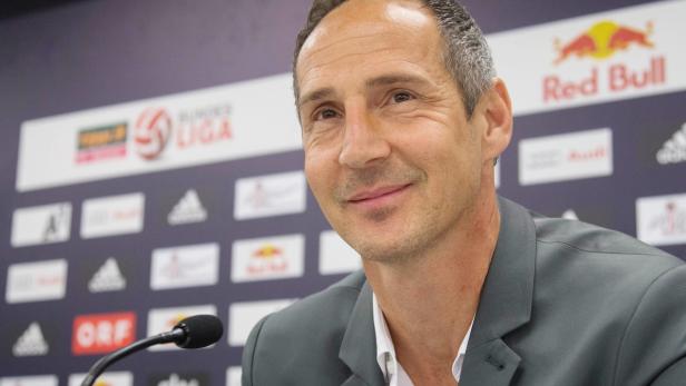Ein Mann in einem grauen Anzug lächelt vor einer Wand mit Logos, darunter Red Bull und Bundesliga.