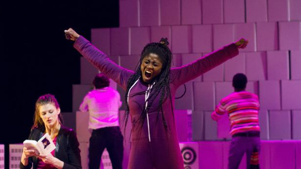 Eine Frau in einem lila Overall steht mit erhobenen Armen auf einer Bühne.