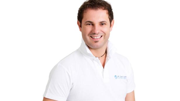 Ein lächelnder Mann mit dunklen Haaren und einem weißen Poloshirt vor einem weißen Hintergrund.