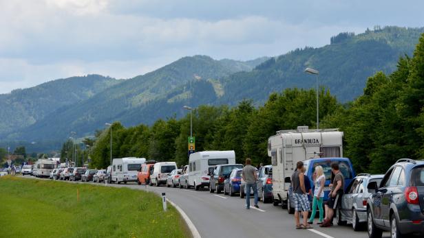 Eine lange Schlange von Autos und Wohnwagen staut sich vor einer bewaldeten Bergkulisse.