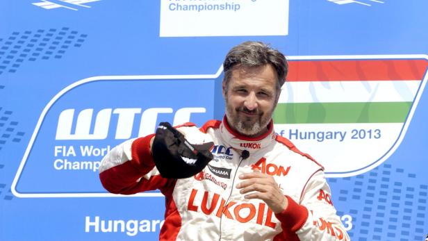 Ein Mann in Rennfahreruniform hält eine Kappe vor einem WTCC-Hintergrund.