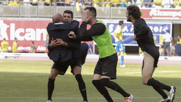 Ein Fußballtrainer umarmt einen Spieler, während andere Spieler unterschiedliche Reaktionen zeigen.