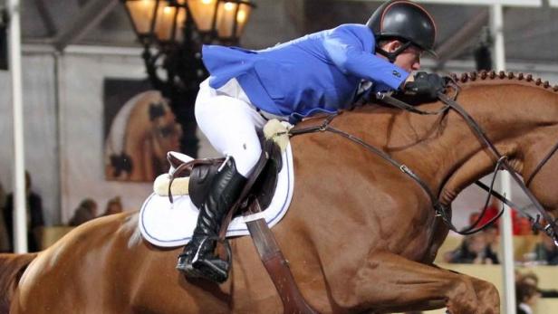 Ein Reiter in blauer Jacke springt mit seinem Pferd über ein Hindernis.